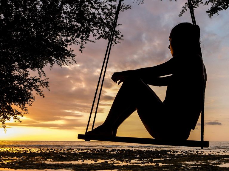Silhouette of a woman sitting on a swing overlooking water illustrating overthinking.